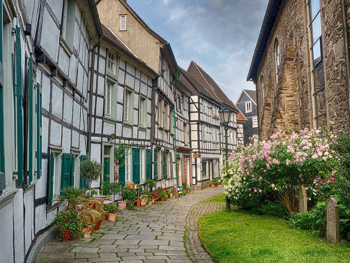 Idyllische, gepflasterte Gasse in einer deutschen Altstadt mit traditionellen Fachwerkhäusern, grünen Fensterläden und blühenden Rosensträuchern.
