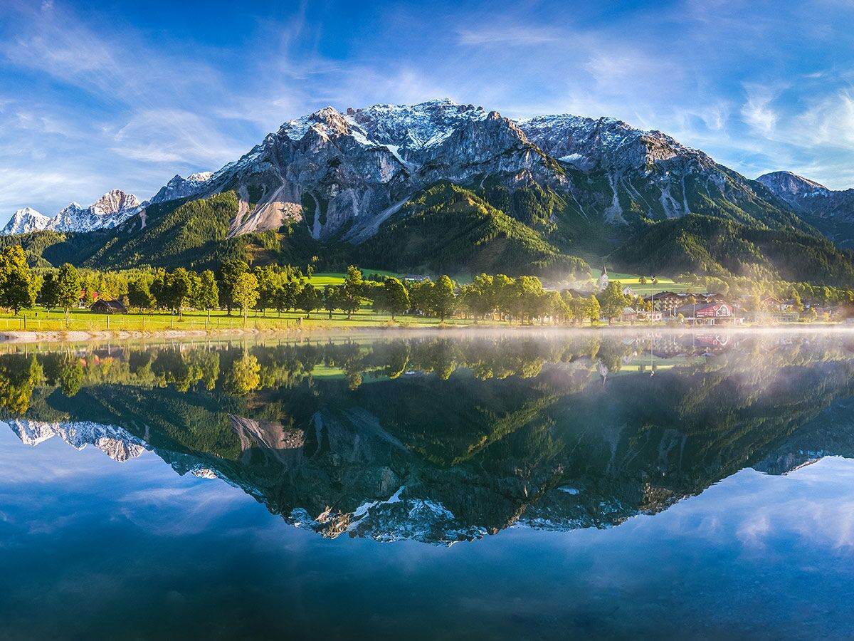 Spiegelung von schneebedeckten Bergen, grünem Wald und einem Dorf im klaren Bergsee bei Sonnenaufgang.