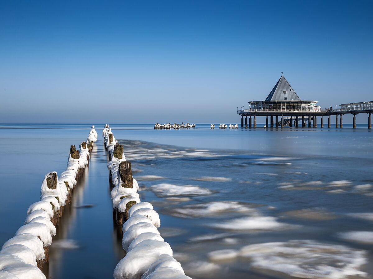 Winterliche Ansicht der Seebrücke Sellin auf Rügen mit eisbedeckten Pfählen im Meer unter blauem Himmel.