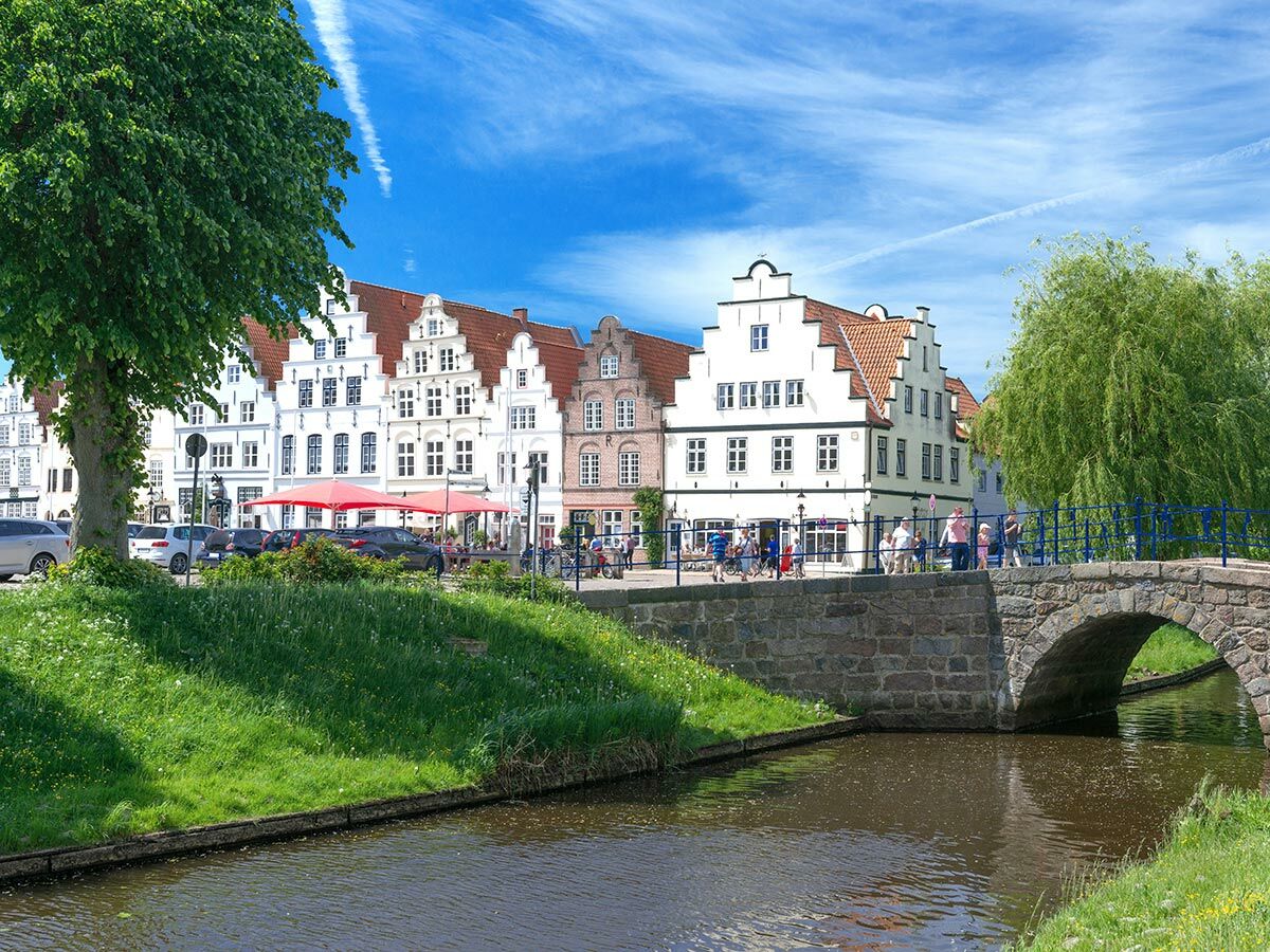 Blick auf eine historische Altstadt mit weißen Giebelhäusern, Kanal, steinerner Brücke, grünen Bäumen und Menschen an einem sonnigen Tag.