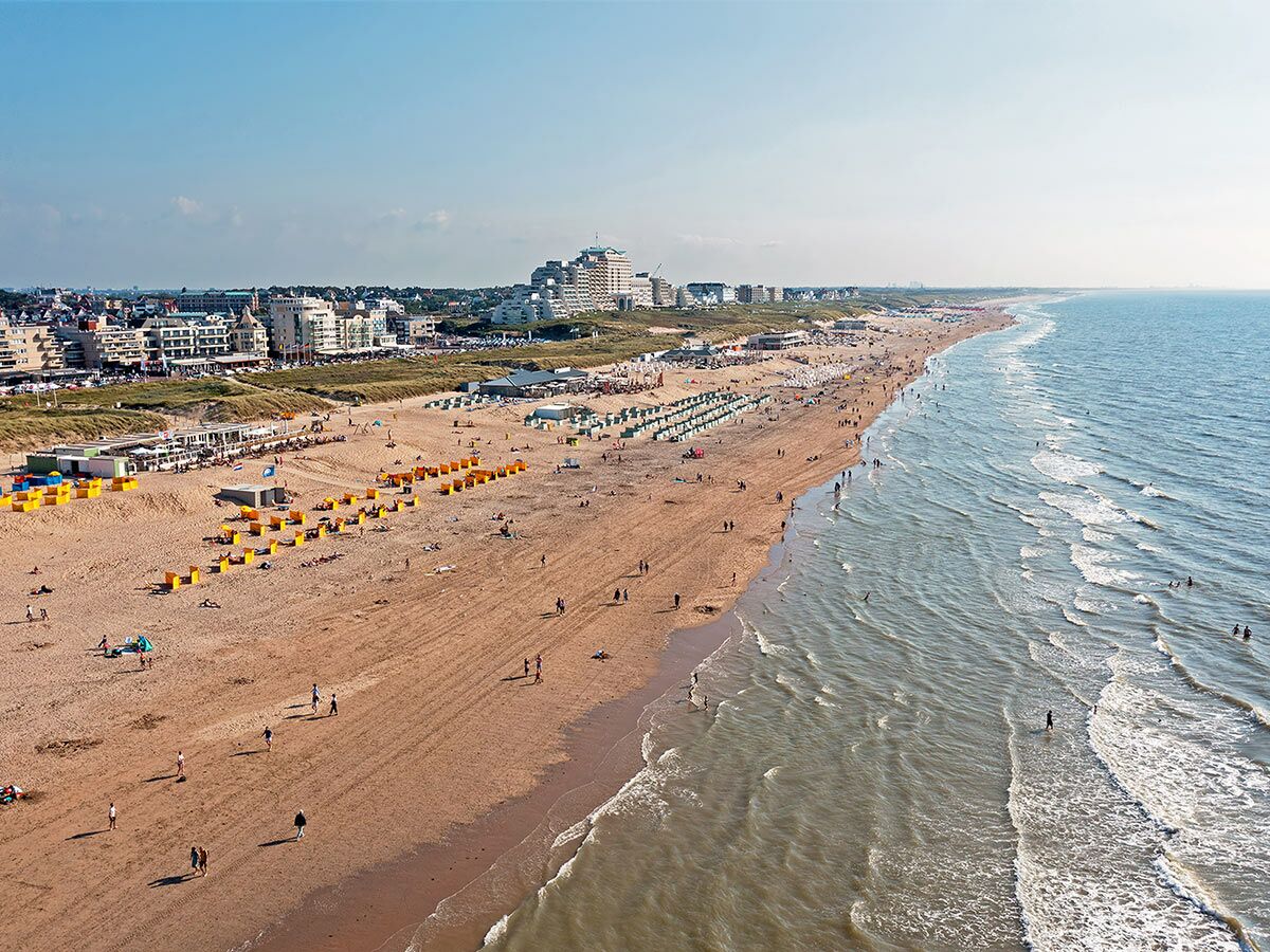 Belebter Sandstrand mit Menschen, die am Meer spazieren und schwimmen, im Hintergrund eine Küstenstadt mit Hotels und Dünen bei sonnigem Wetter.