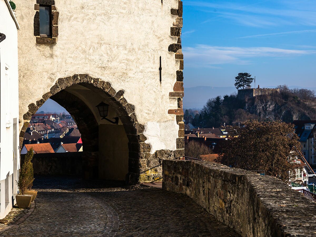 Ein alter Steintorbogen in einer Altstadt, der einen Blick auf rote Dächer und eine Burgruine auf einem Hügel freigibt.