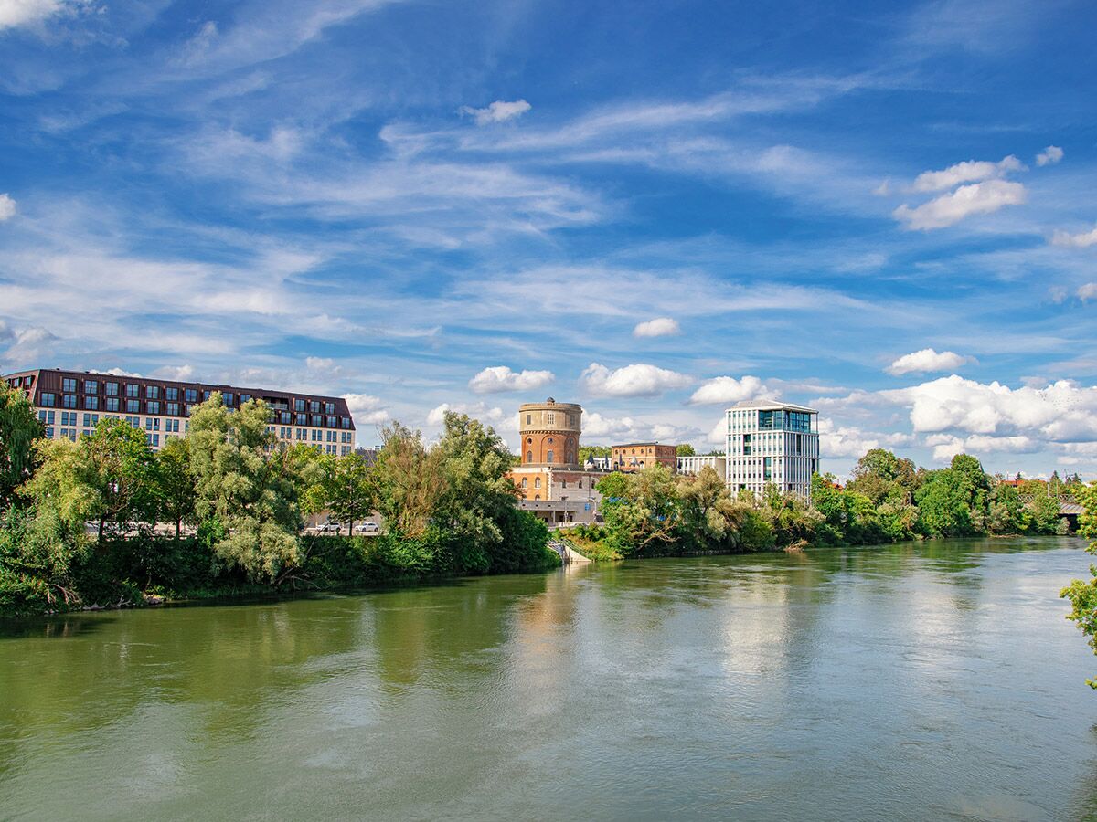 Stadtansicht Ingolstadt am Fluss: Grüne Uferbäume, moderner Gebäudekomplex, historischer Wasserturm unter blauem Himmel mit Wolken.