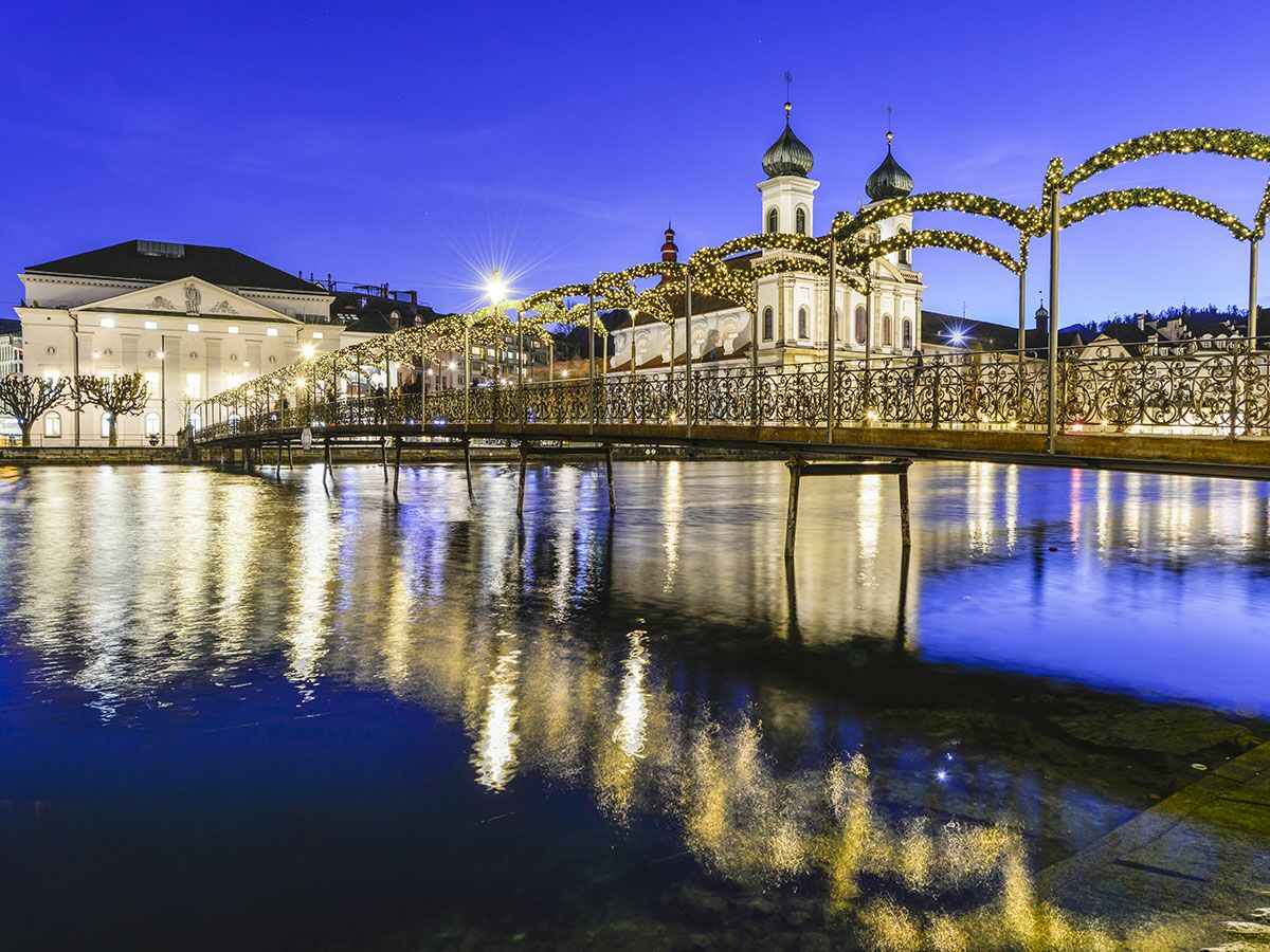 Luzerner Jesuitenkirche und geschmückte Brücke bei Blaue Stunde, mit Lichtreflexionen im Fluss Reuss.