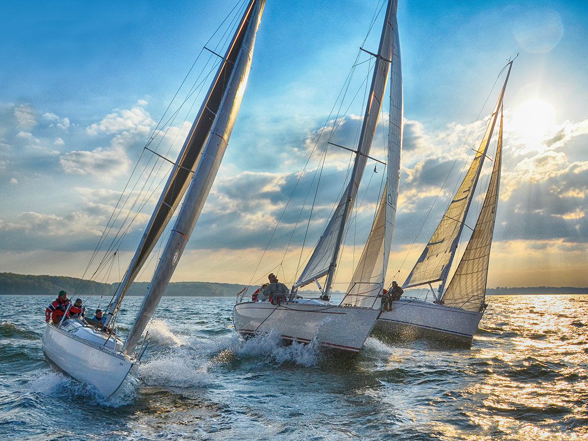 Drei Segelboote rasen bei goldenem Sonnenuntergang mit spritzendem Wasser über das Meer.