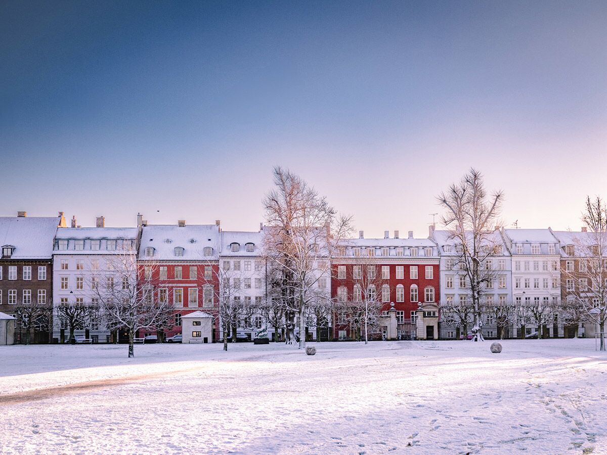 Winterliche Stadtansicht mit verschneiten Gebäuden, kahlen Bäumen und einem schneebedeckten Park bei Sonnenaufgang oder -untergang.