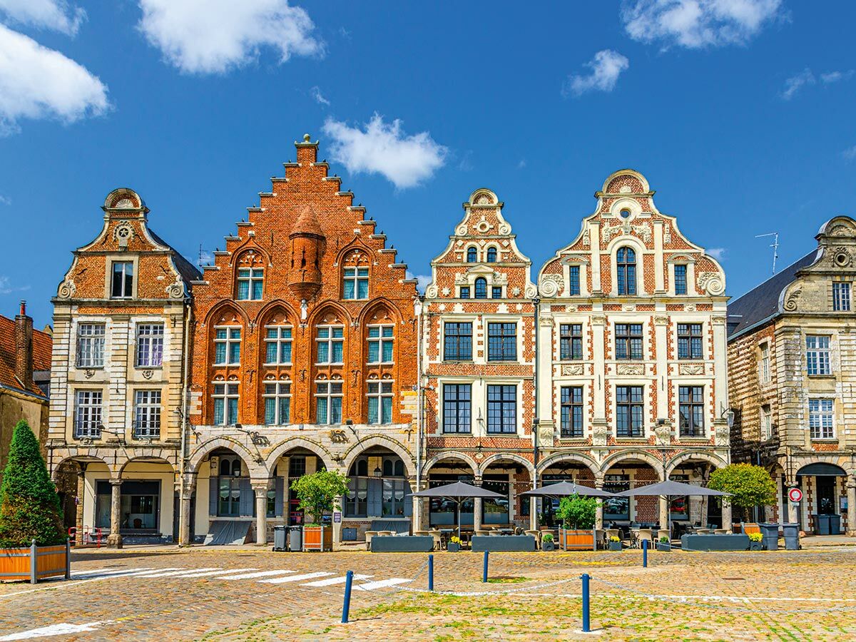 Reihe historischer Giebelhäuser aus Backstein und Stein an einem sonnigen Marktplatz mit Kopfsteinpflaster und Cafés.