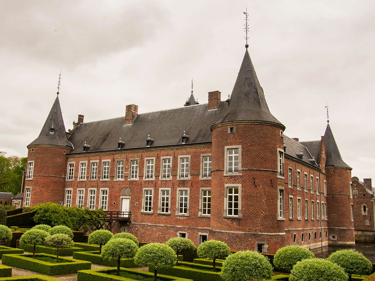Historisches Backstein-Wasserschloss mit spitzen Türmen, Wassergraben und gepflegten Gärten unter bewölktem Himmel.