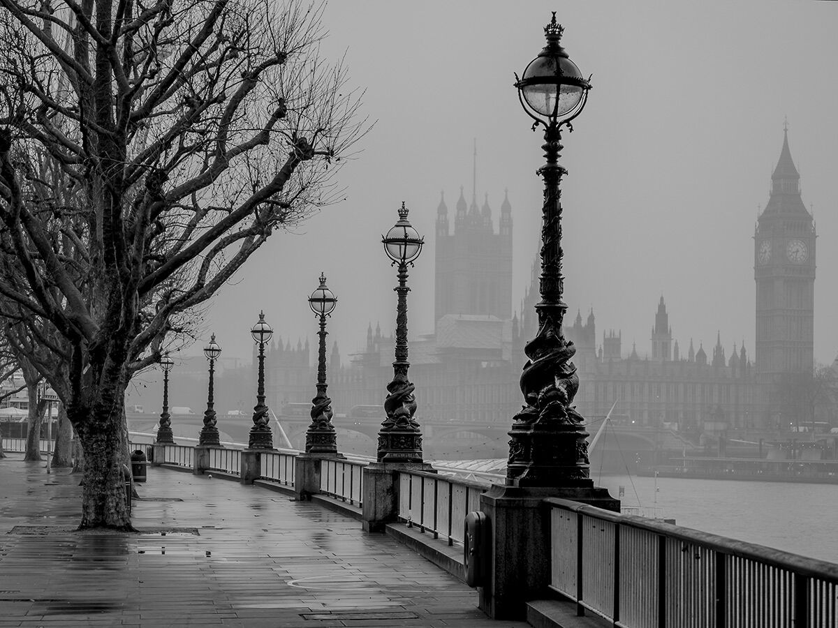 Schwarz-Weiß-Bild von London im Nebel mit Blick auf die Themse, die Houses of Parliament und Big Ben hinter historischen Laternen und Bäumen.