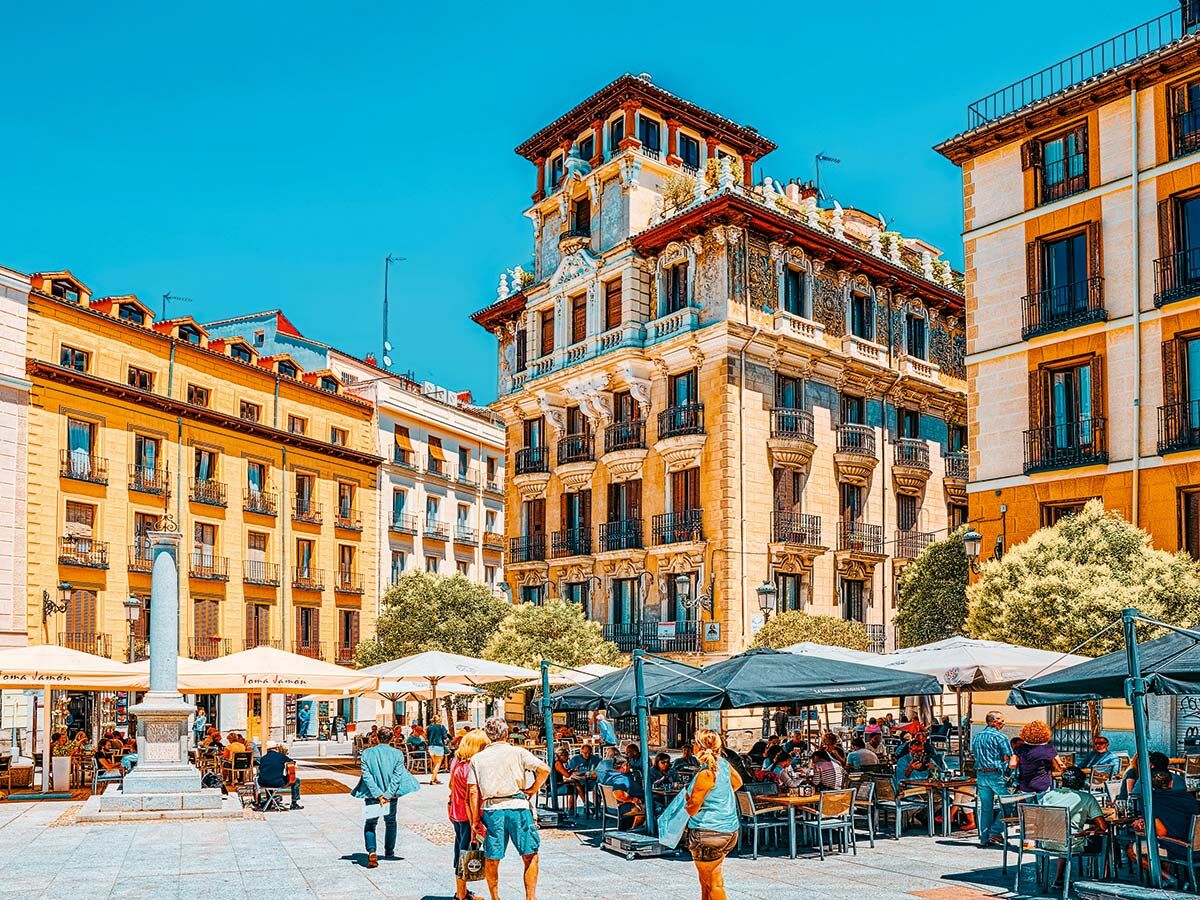 Belebter städtischer Platz in Madrid mit historischen Gebäuden, Menschen auf Sonnenterrassen und blauem Himmel.