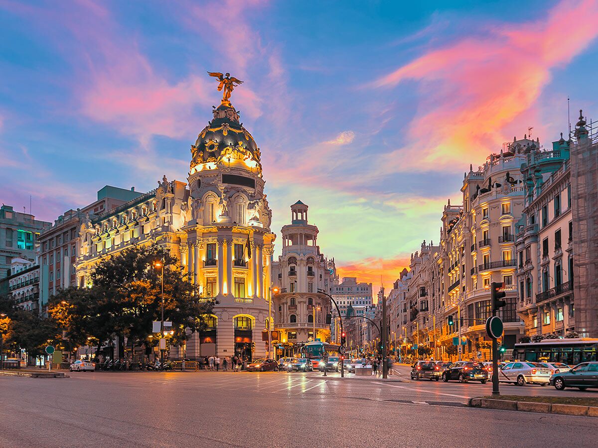 Das ikonische Metrópolis Gebäude in Madrid bei Sonnenuntergang mit pinken Wolken, beleuchteter Fassade, Verkehr und Fußgängern.