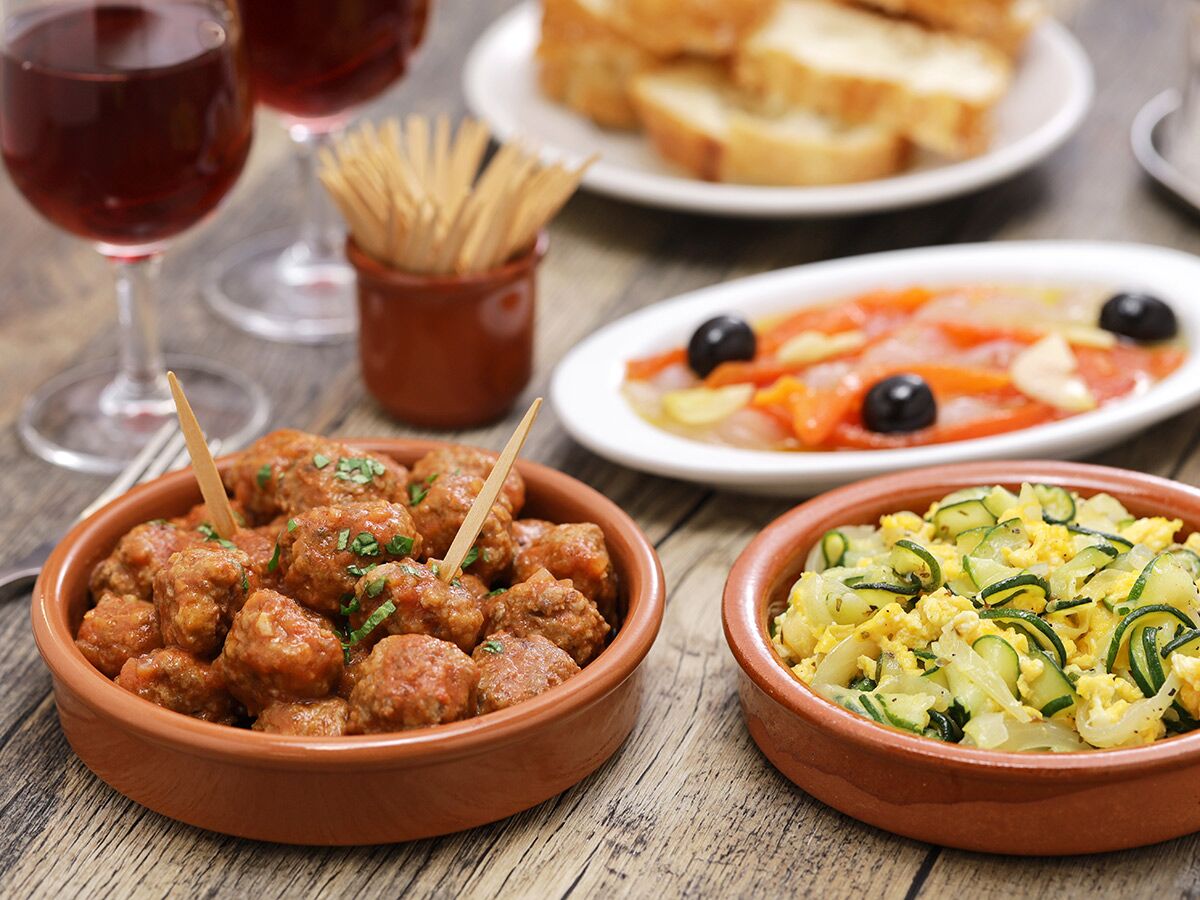 Teller mit spanischen Tapas: Albóndigas, Zucchini-Rührei, Paprika mit Oliven; dazu Rotwein und Brot auf Holztisch.
