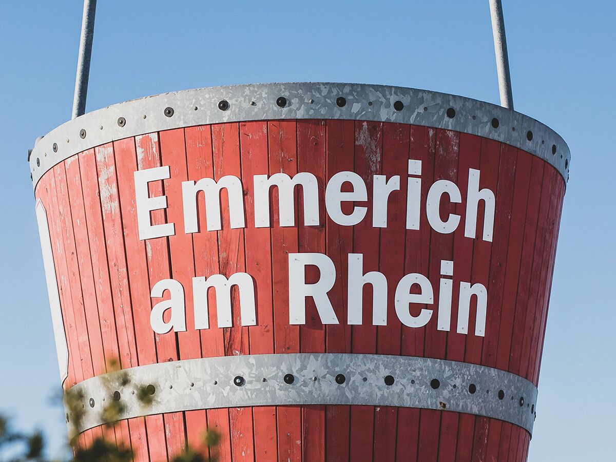 Großer roter Holzfass-Wegweiser mit weißer Aufschrift "Emmerich am Rhein" vor blauem Himmel.