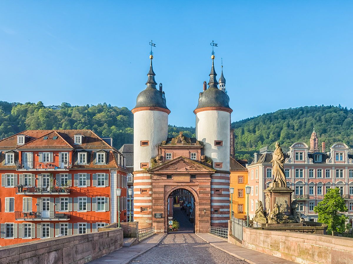Heidelberg: Historisches Brückentor der Alten Brücke mit Türmen, Statue und farbigen Gebäuden vor grünen Hügeln.