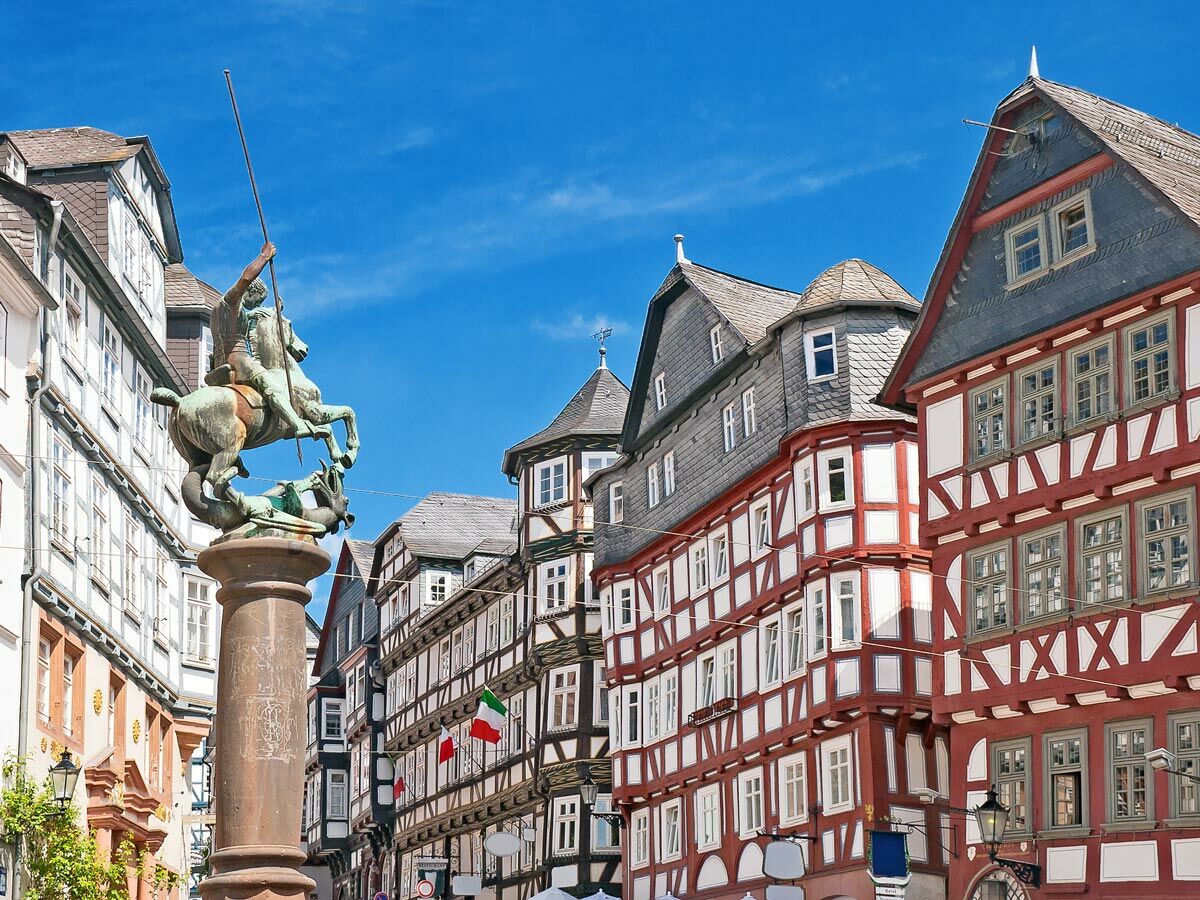 Bronzestatue von St. Georg auf Pferd im Kampf mit Drachen, umgeben von Fachwerkhäusern unter blauem Himmel.