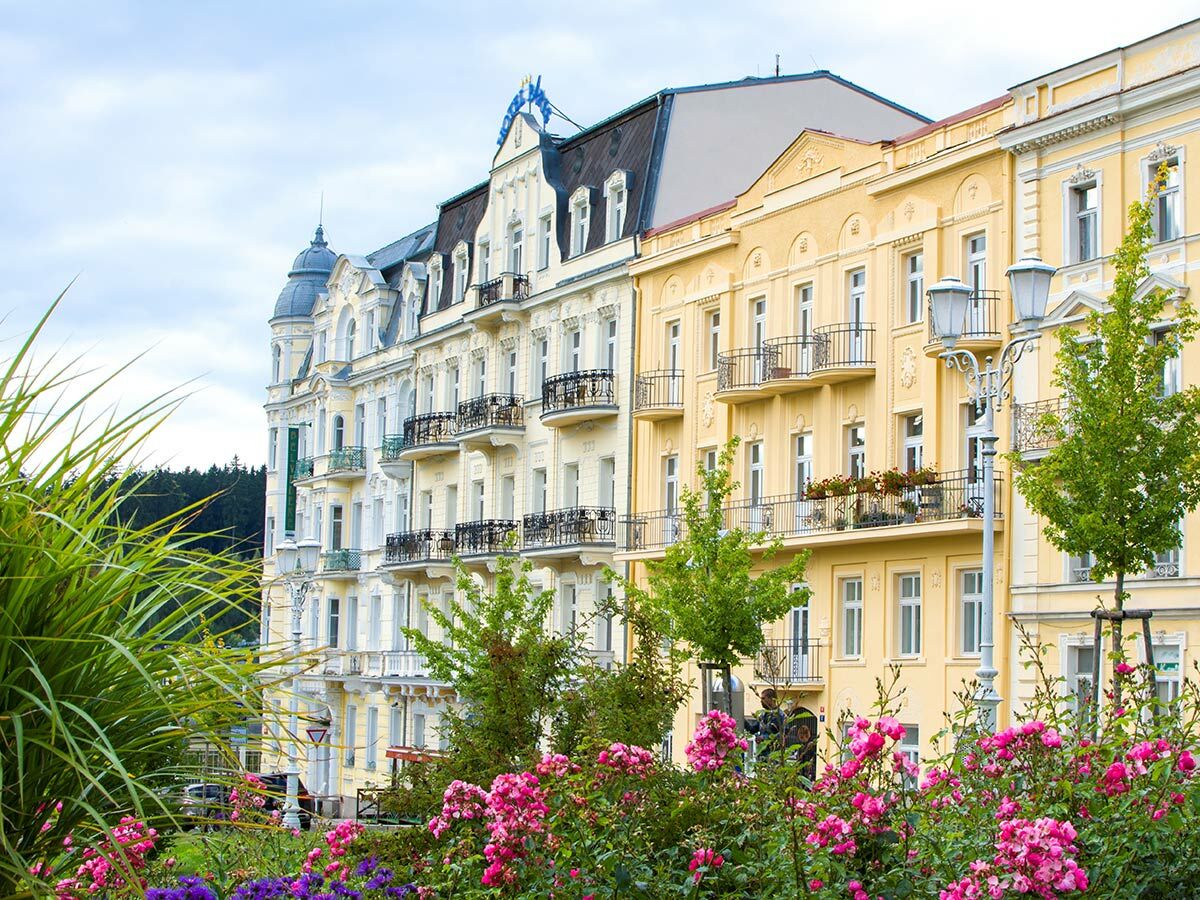 Reihe historischer Gebäude mit Balkonen und aufwendigen Fassaden. Im Vordergrund blühende pinke und lila Blumen und grüne Pflanzen.