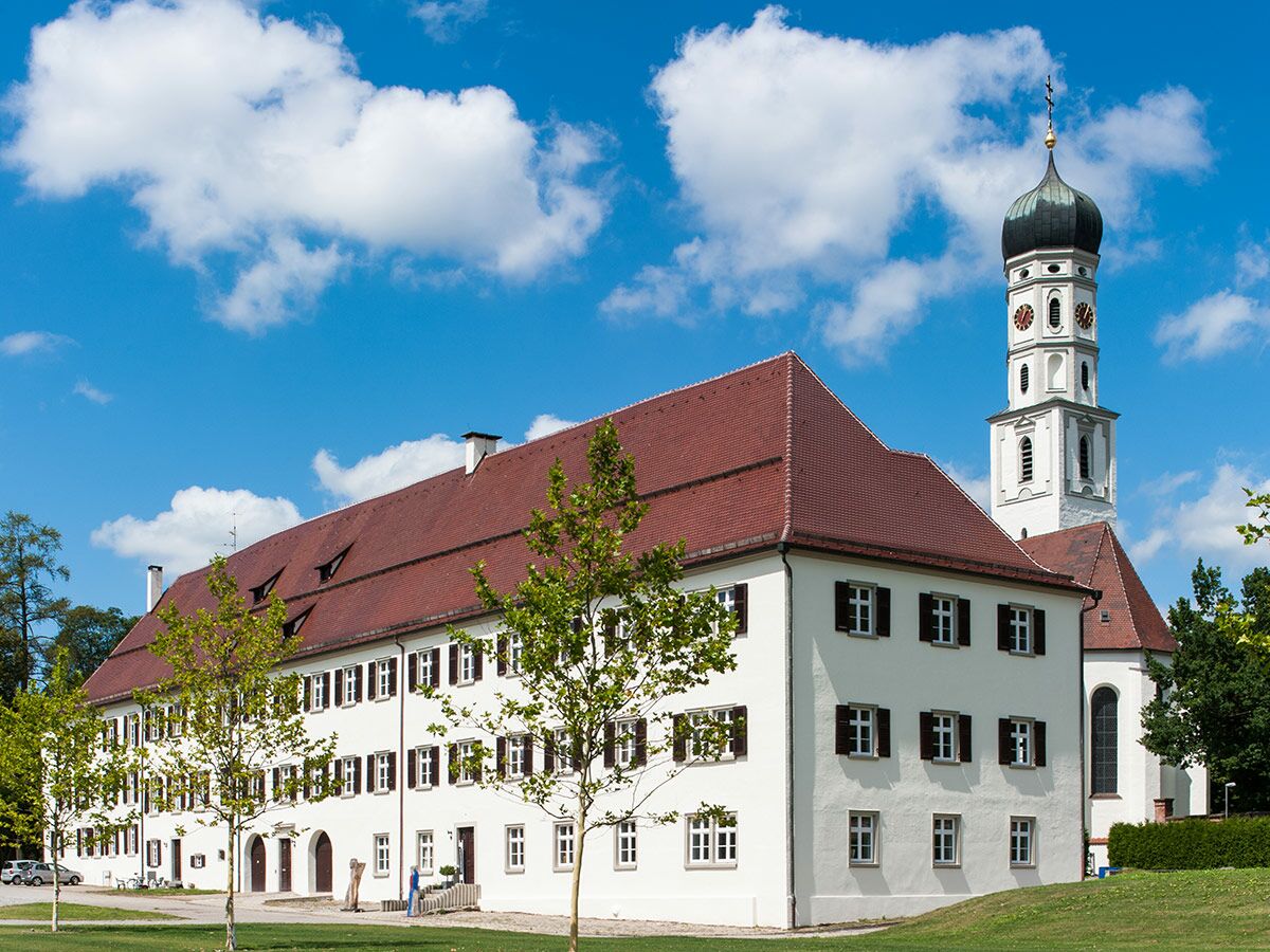 Weißes, historisches Klostergebäude mit rotem Dach und hohem barocken Kirchturm mit Kuppel unter blauem Himmel mit Wolken.