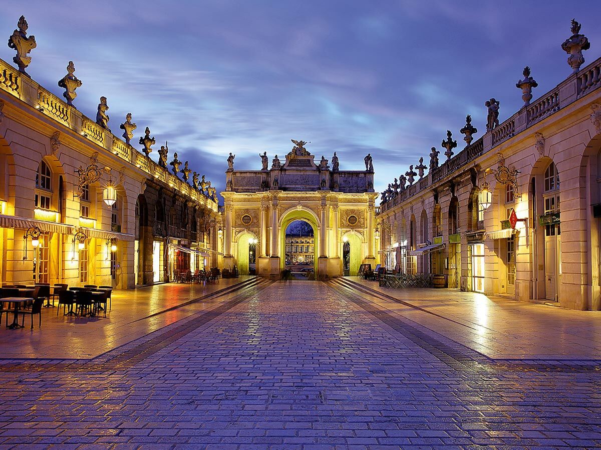 Historische Porte Héré und Place Stanislas in Nancy bei Abenddämmerung mit warm beleuchteten Fassaden und Geschäften.