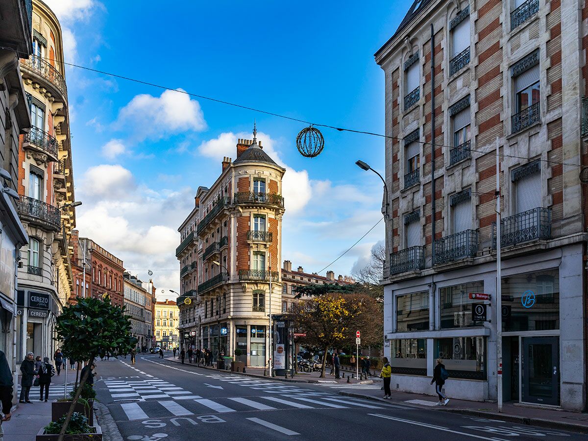 Belebte europäische Stadtstraße mit historischen Gebäuden, Fußgängern auf dem Bürgersteig und Zebrastreifen bei blauem Himmel und Wolken.