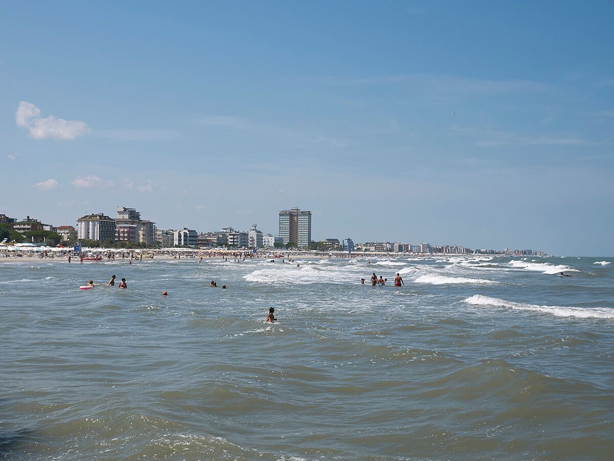 Sonniger Sommertag am Strand: Viele Menschen schwimmen im Meer vor einer belebten Küstenlinie mit Hotels und Gebäuden.