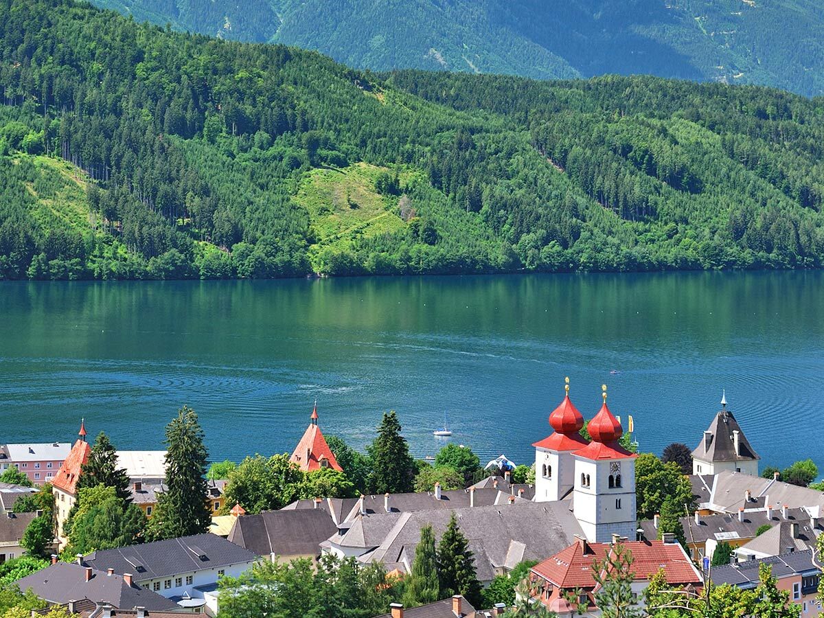 Idyllische Stadt mit roten Zwiebeltürmen am See, umgeben von grünen Wäldern und Bergen.