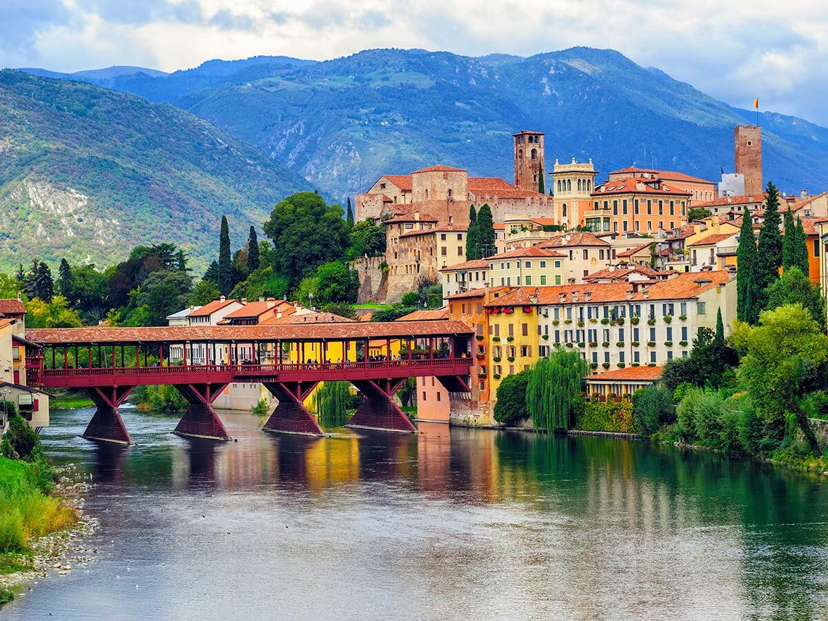 Malerisches Bassano del Grappa mit der historischen Ponte Vecchio über dem Brenta-Fluss, umgeben von bunten Häusern und grünen Bergen.