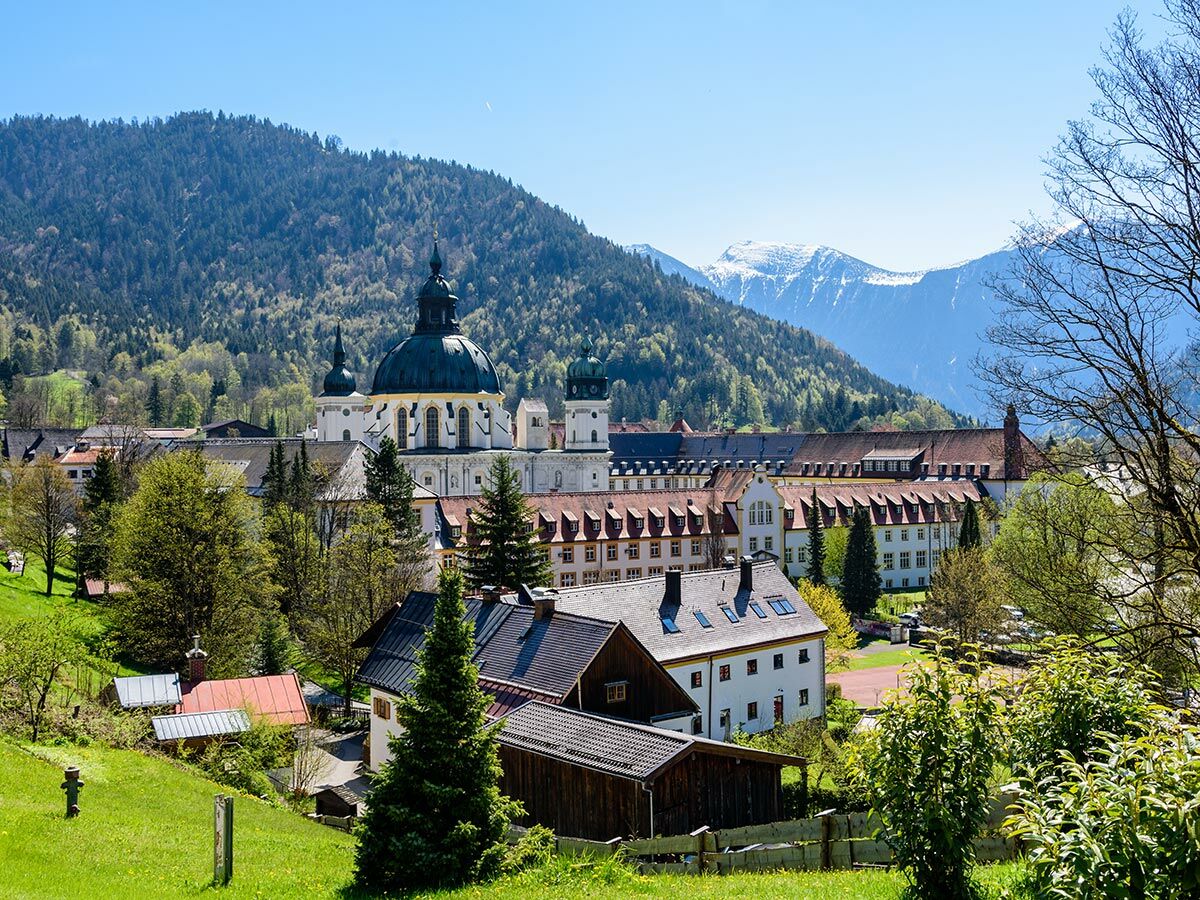 Panoramablick auf Kloster Ettal in den bayerischen Alpen, umgeben von grünen Hügeln, Bäumen und schneebedeckten Bergen.