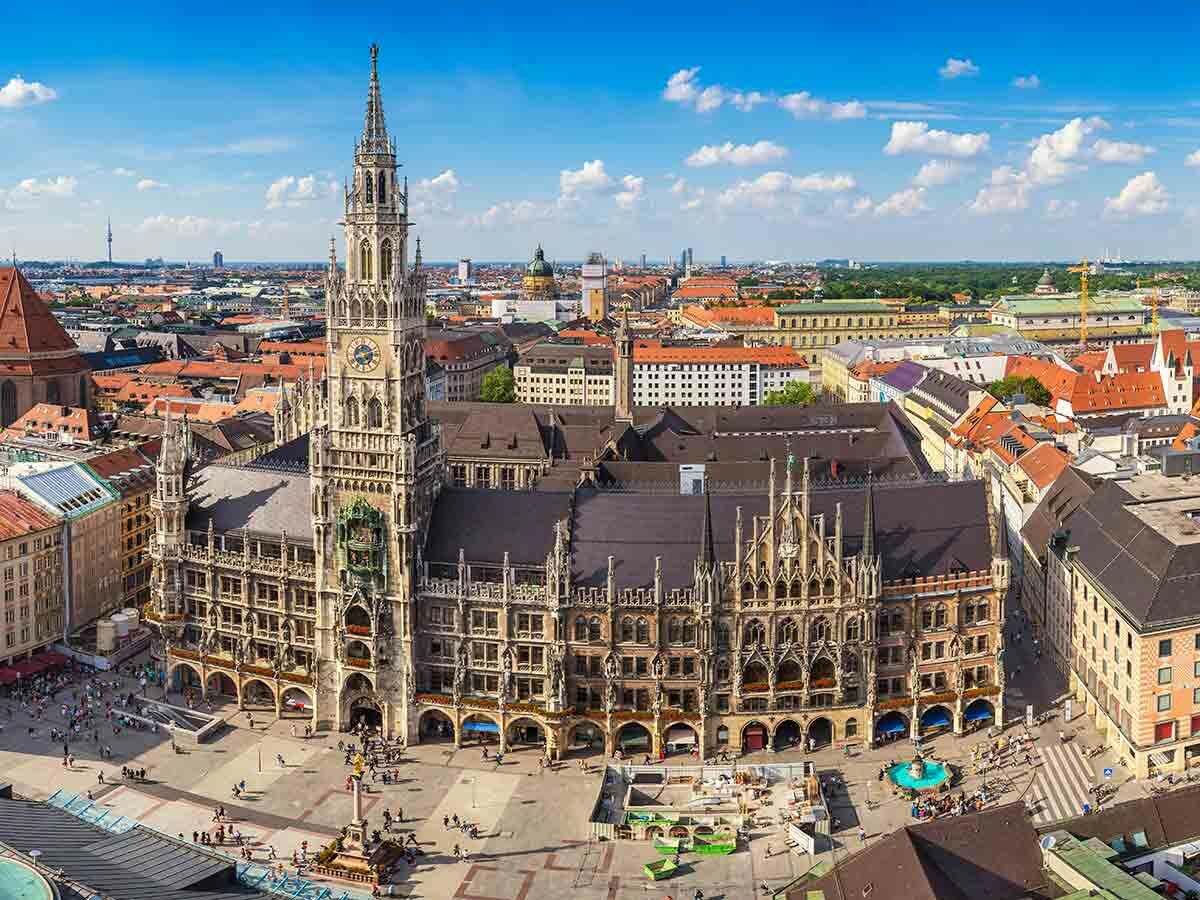 Luftaufnahme des Neuen Rathauses am Marienplatz in München mit Menschenmenge und umliegender Stadt unter blauem Himmel.