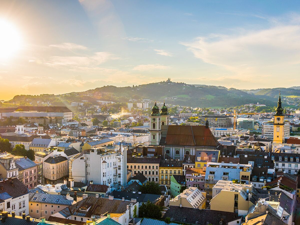 Panoramablick auf Linz bei Sonnenuntergang. Historische Gebäude, Kirchen, Wohnhäuser und grüne Hügel am Horizont.