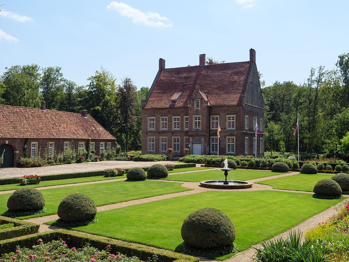 Historisches Backstein-Herrenhaus mit rotem Ziegeldach, umgeben von einem gepflegten Garten mit Brunnen, Rasen und Hecken.
