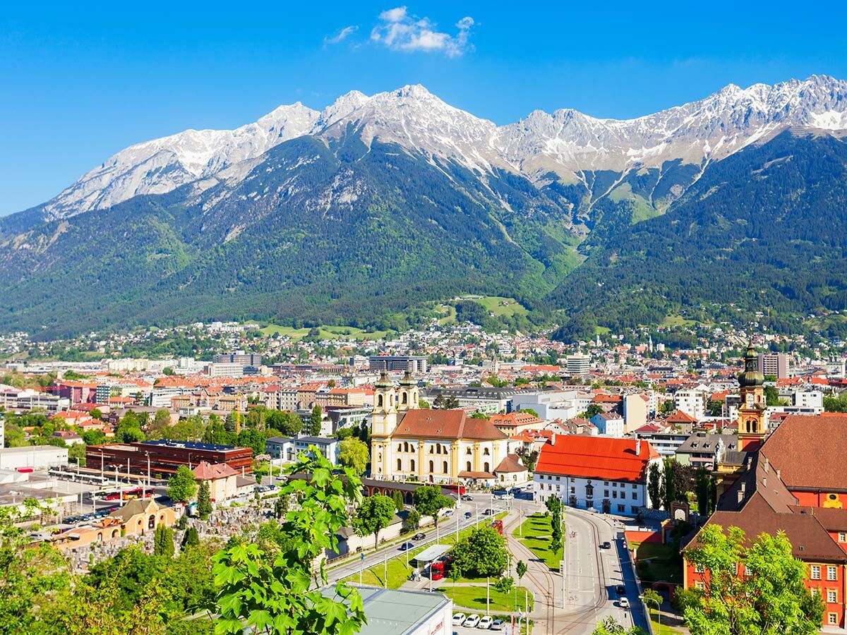 Panoramablick auf die Stadt Innsbruck, umgeben von schneebedeckten Alpen und grünen Hügeln unter blauem Himmel.