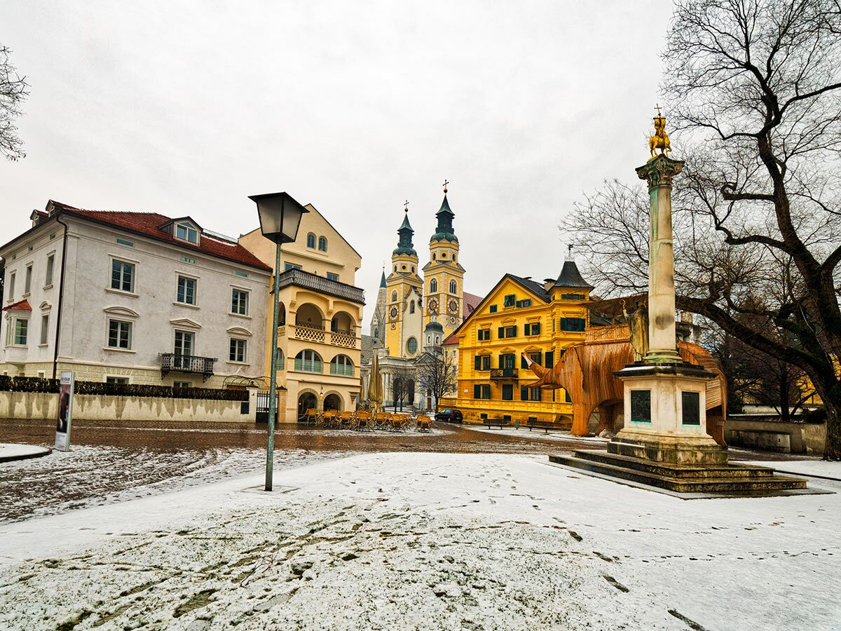 Schneebedeckter Domplatz Brixen mit Kathedrale, historischen Gebäuden, Soliman-Säule und Holz-Elefanten unter bewölktem Himmel im Winter.
