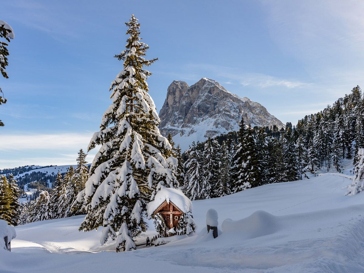 Idyllische, verschneite Winterlandschaft in den Alpen mit großen Fichtenbäumen, einem hölzernen Kreuzwegschrein und majestätischen Bergen.
