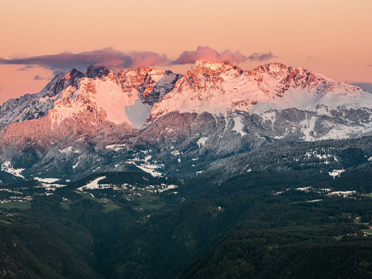 Verschneite Berge leuchten rosa im Sonnenuntergang über einem bewaldeten Tal mit kleinen Dörfern.