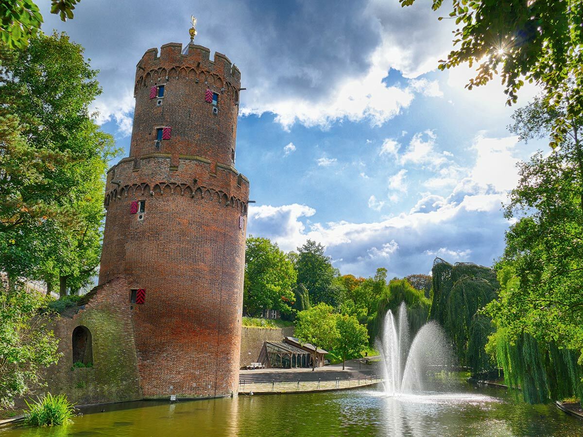 Runder Ziegelsteinturm am Wasser mit plätschernden Brunnen und grünen Bäumen unter sonnigem, bewölktem Himmel.