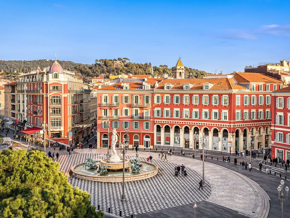 Luftaufnahme des Place Masséna in Nizza mit Apollo-Brunnen, roten Gebäuden, grünen Fensterläden und Menschen.