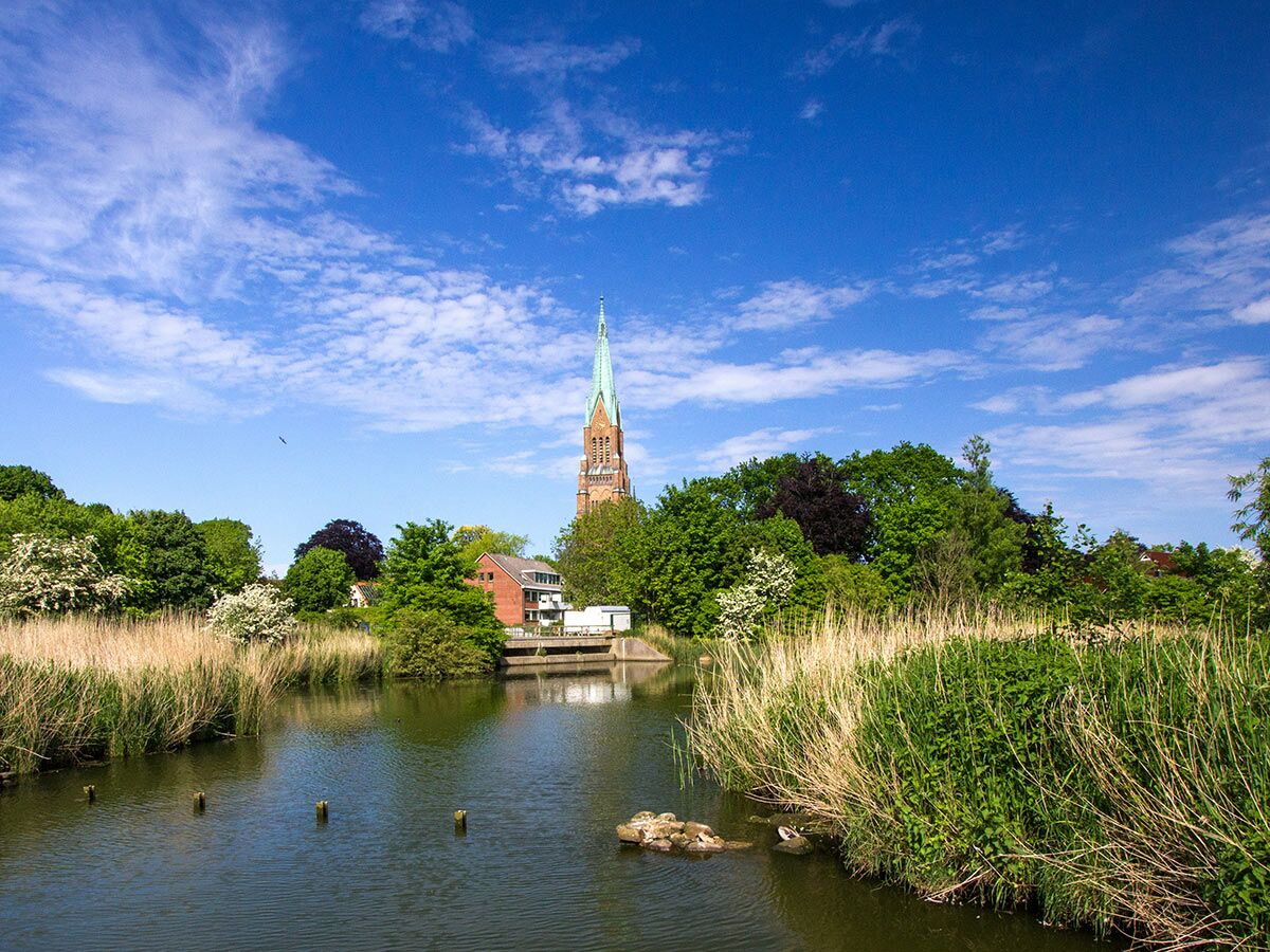 Backstein-Kirchturm mit grünem Dach an einem Flussufer, umgeben von Schilf, Bäumen und Häusern unter blauem Himmel.