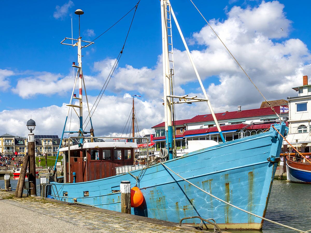 Ein traditionelles blaues Fischerboot liegt an einem sonnigen Tag am Kai eines belebten Hafens mit Gebäuden und Masten im Hintergrund.