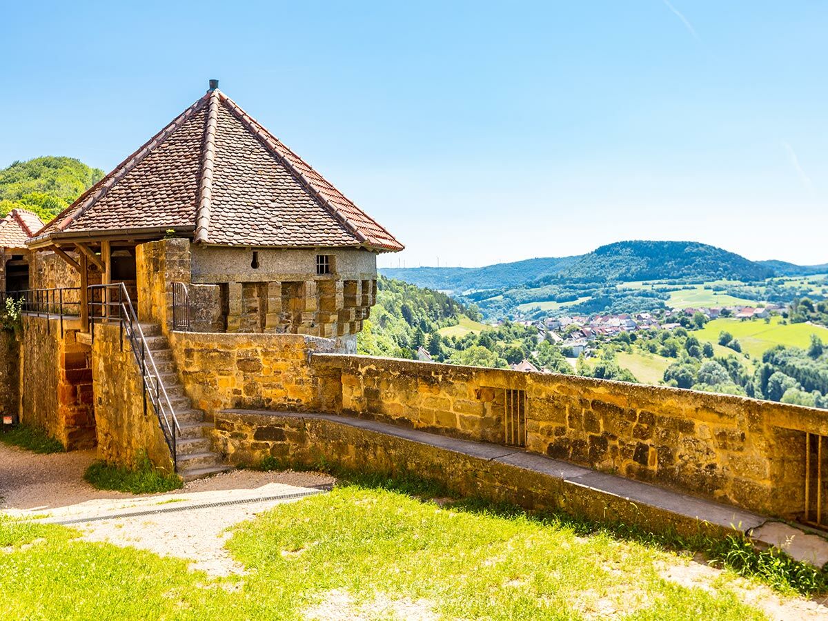 Historischer Burgturm mit Treppen und Steinmauer; Blick auf ein grünes Tal mit Dorf, Wäldern und Hügeln unter blauem Himmel.