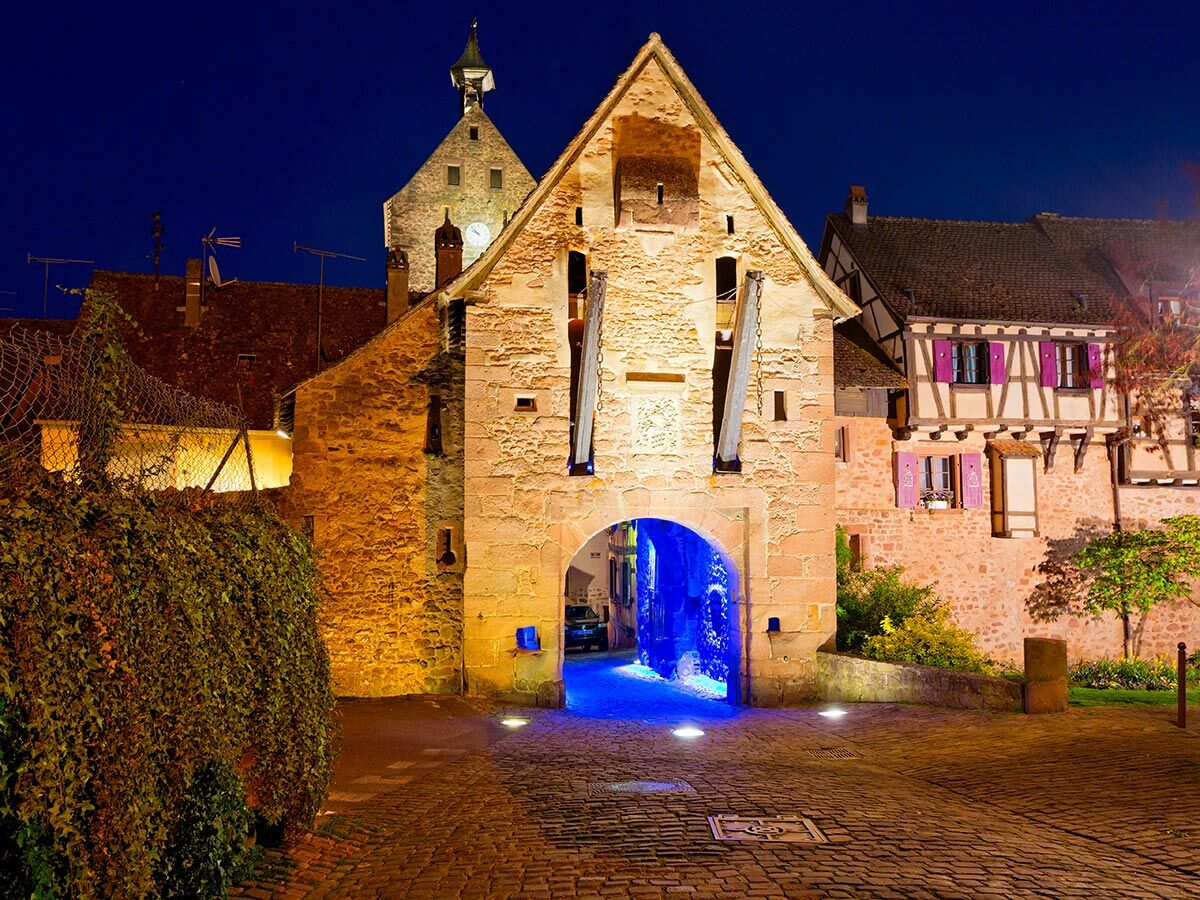 Historisches Steintor mit blauer Bogenbeleuchtung und Fachwerkhäusern in einem europäischen Dorf bei Nacht, Kopfsteinpflaster.