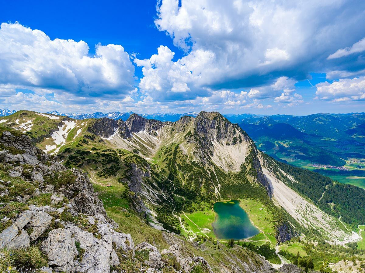 Atemberaubendes Bergpanorama mit tiefblauem See, grünen Wiesen und schneebedeckten Gipfeln unter blauem Himmel mit Wolken.