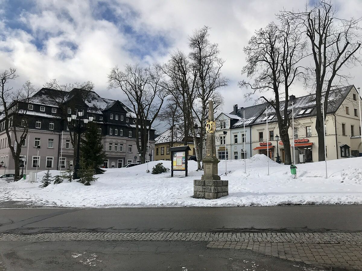Winterliche Szene eines verschneiten Stadtplatzes mit historischen Gebäuden, kahlen Bäumen und einem Denkmal im Harz.