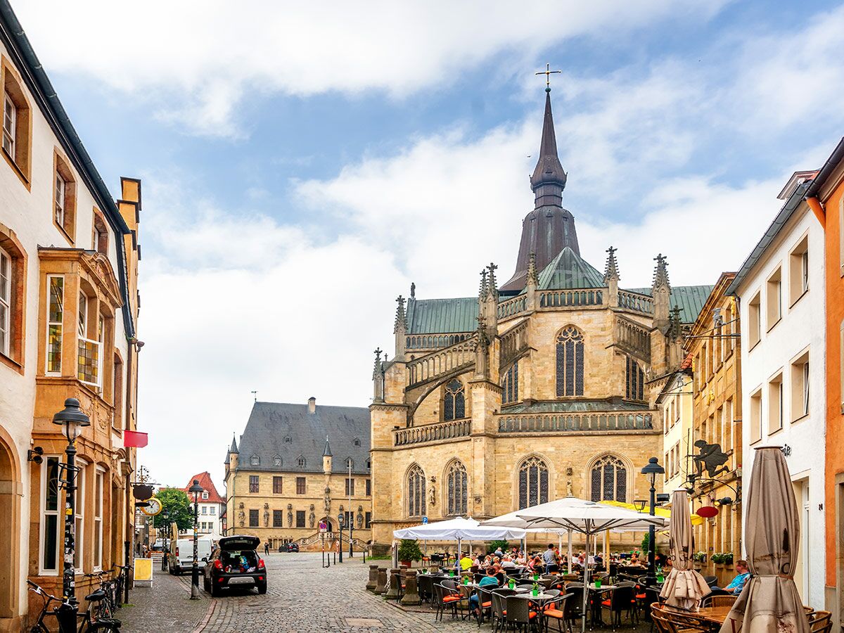 Historischer Domplatz in Osnabrück mit St. Peter Kathedrale, belebten Cafés und traditionellen Gebäuden unter blauem Himmel.