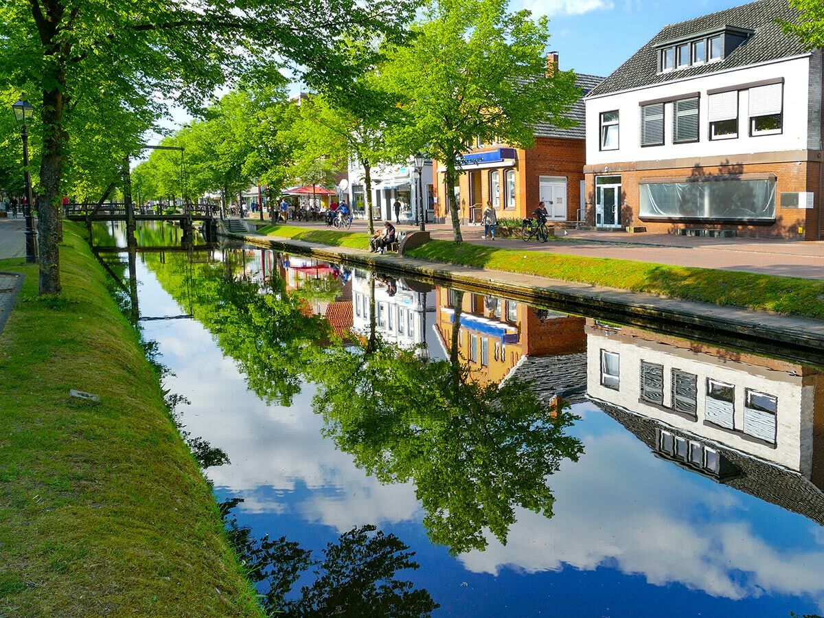 Idyllischer Kanal in einer Stadt mit grünen Bäumen und Gebäuden, die sich im blauen Wasser spiegeln, Menschen am Ufer.