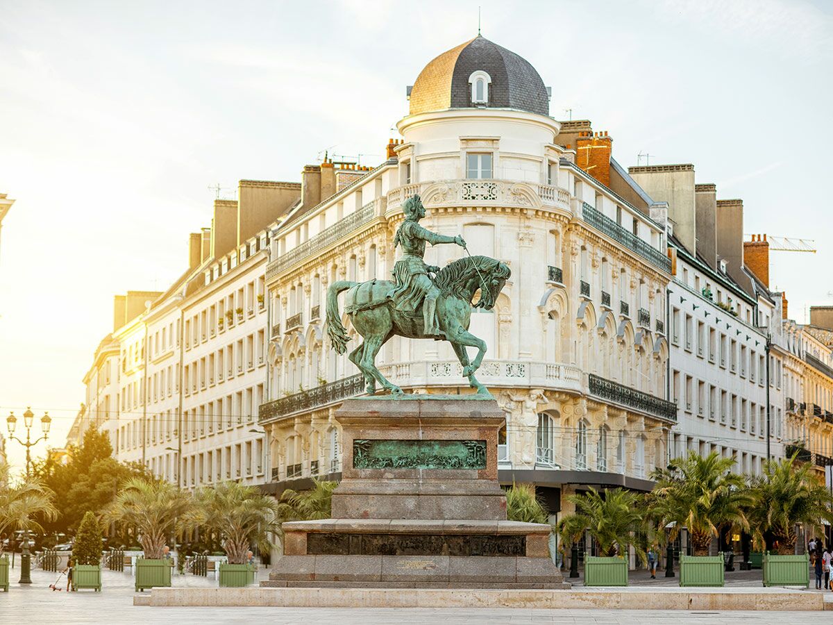 Bronze-Reiterstatue Jeanne d'Arc in Orléans auf sonnigem Stadtplatz mit historischen Gebäuden und Palmen im Hintergrund.