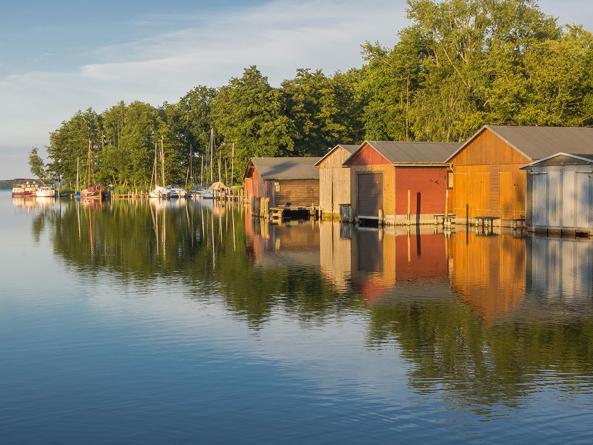 Bunte Bootshäuser und Segelboote spiegeln sich im ruhigen Wasser eines Sees, umgeben von grünen Bäumen unter blauem Himmel.