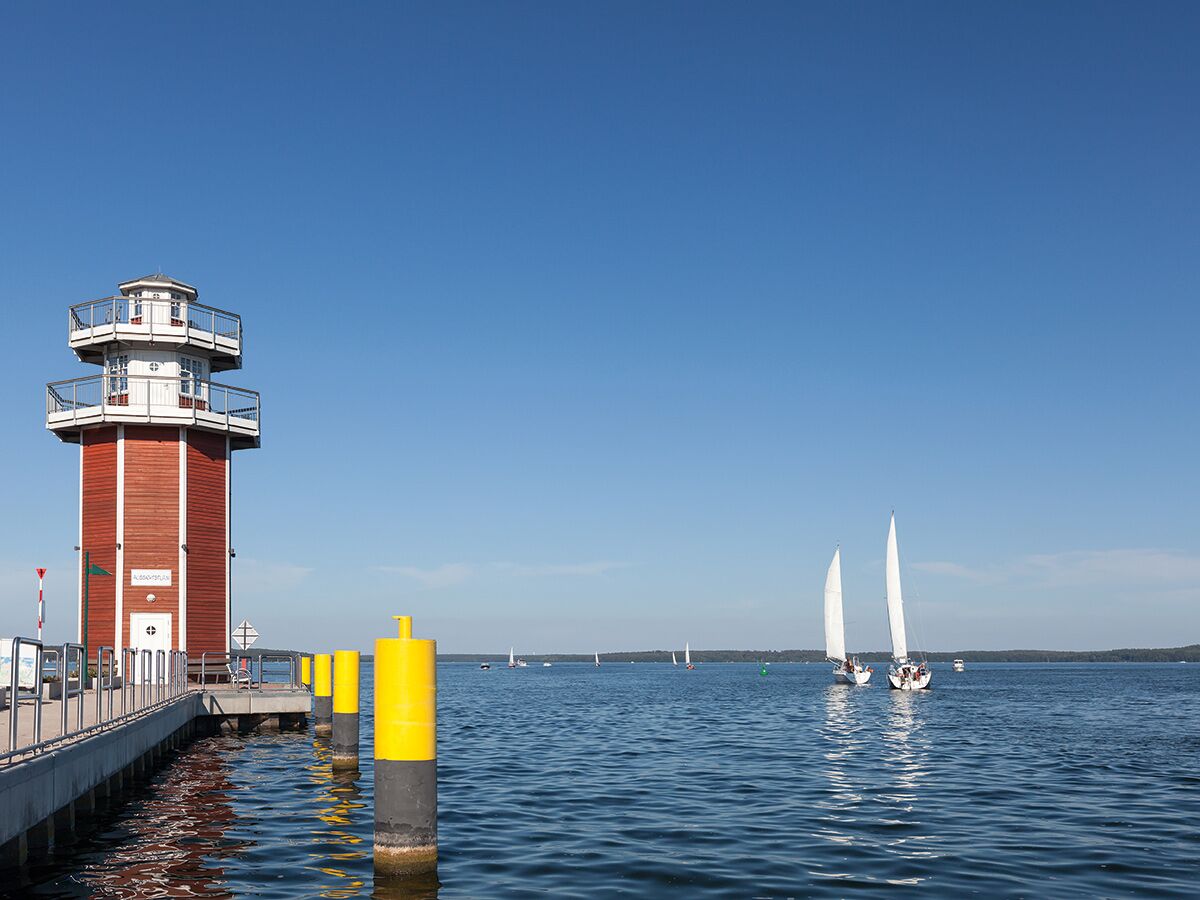 Roter und weißer Leuchtturm an einem Pier auf einem blauen See, im Hintergrund segeln Boote unter klarem Himmel.