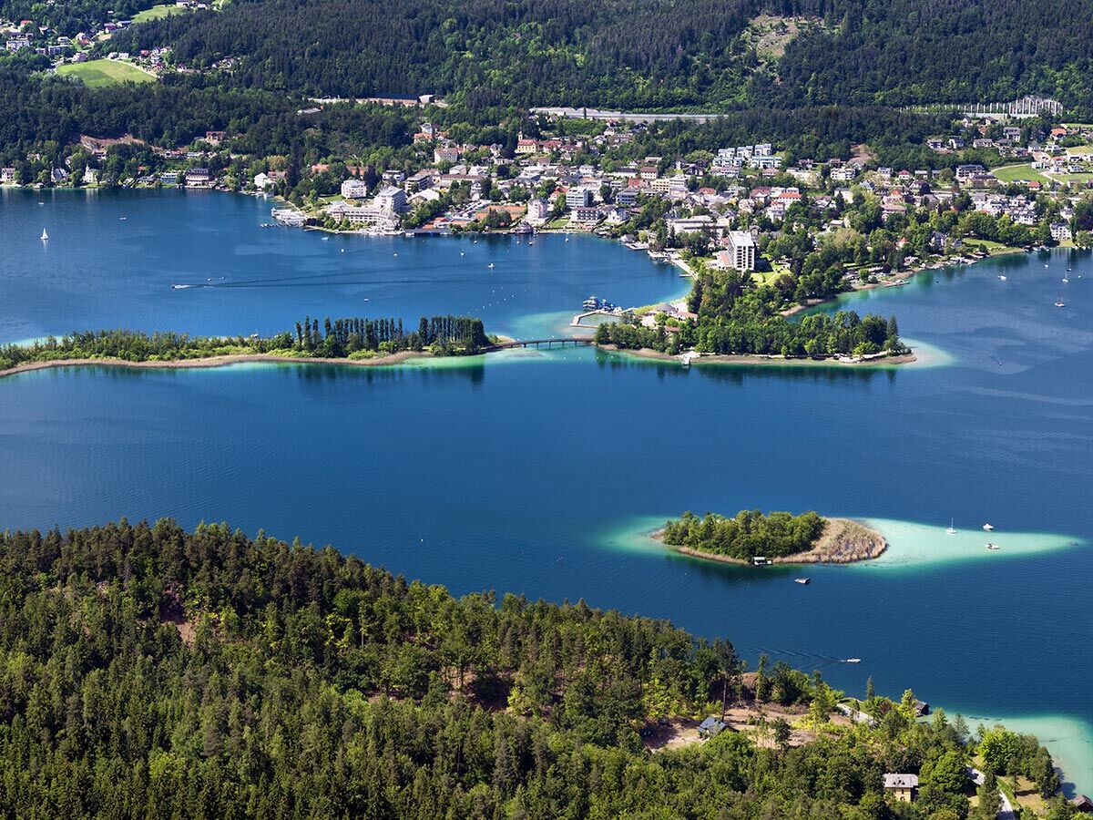 Luftbild eines blauen Sees mit grünen Inseln, verbunden durch Brücken, einer Küstenstadt und bewaldeten Hügeln.