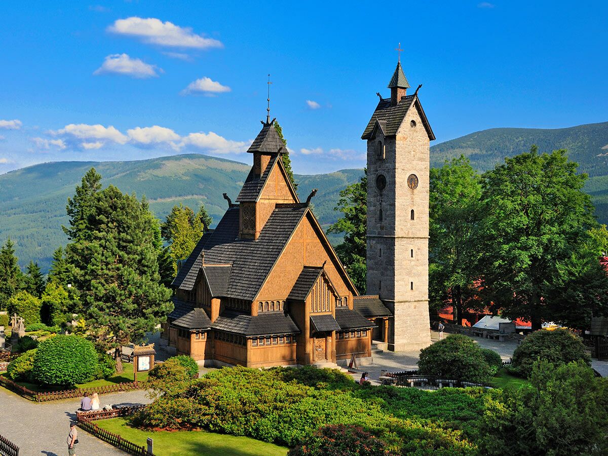 Historische Vang Stabkirche aus Holz mit Steinturm in Karpacz, Polen. Umgeben von grünen Bergen und Bäumen unter blauem Himmel.