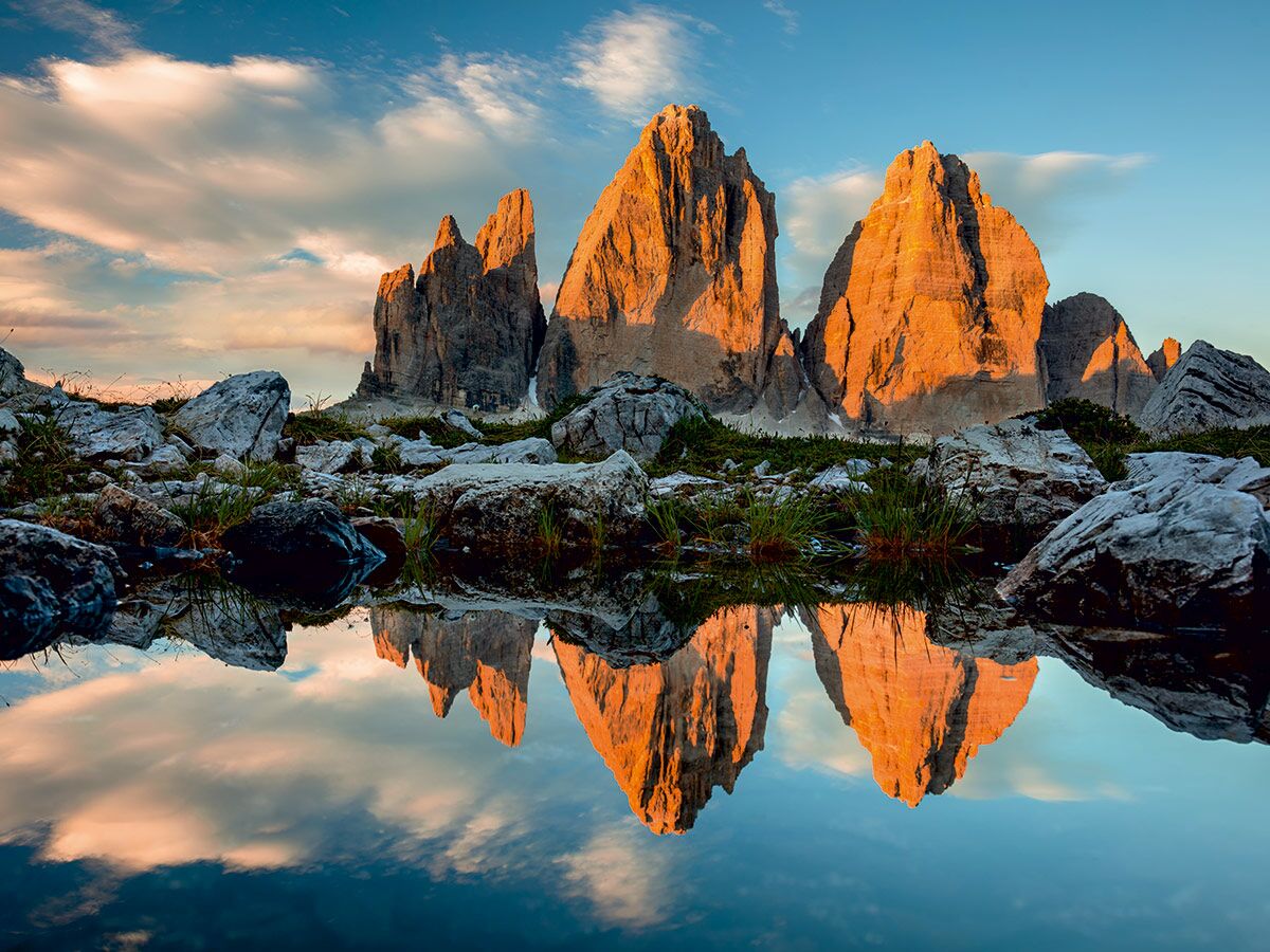 Majestätische Drei Zinnen in den Dolomiten bei Sonnenaufgang, orange beleuchtet, spiegeln sich klar im stillen Bergsee zwischen Felsen.